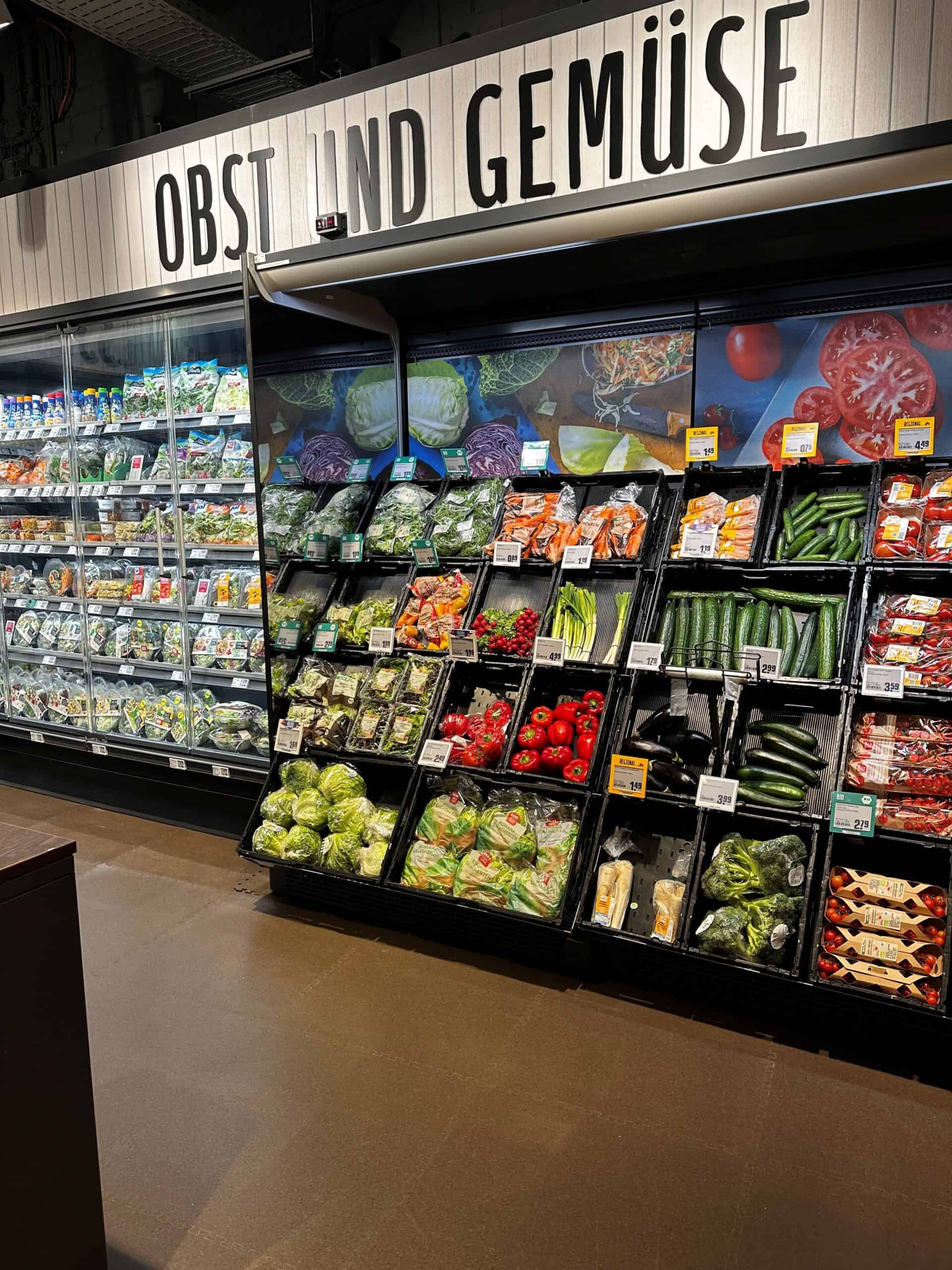 Frisse groenten en fruit in een supermarkt, inclusief tomaten, komkommers, paprika's en sla, gepresenteerd in een overzichtelijke groenteschap met prijskaartjes.