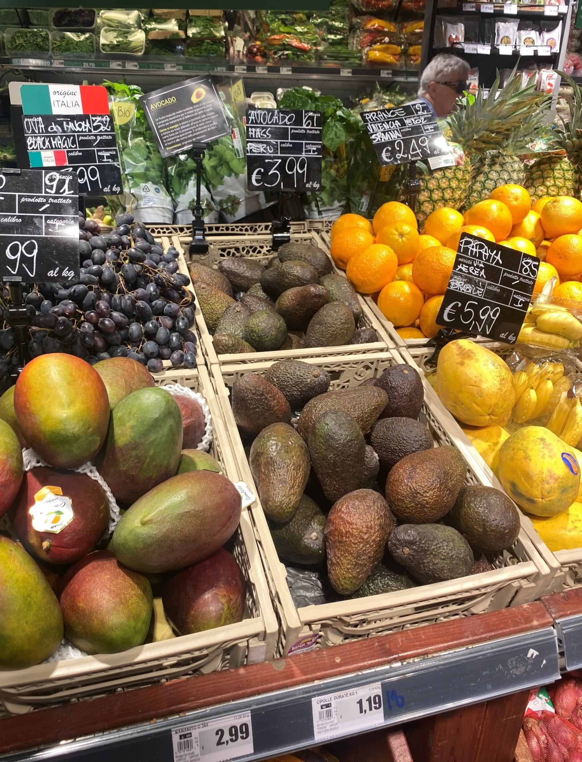 Verse biologische avocado's en andere exotische vruchten in een supermarkt. Geprijsd tussen €2,99 en €3,99 per kilogram, met kleurrijke fruitsoorten zoals mango's, perziken, en citrusvruchten.