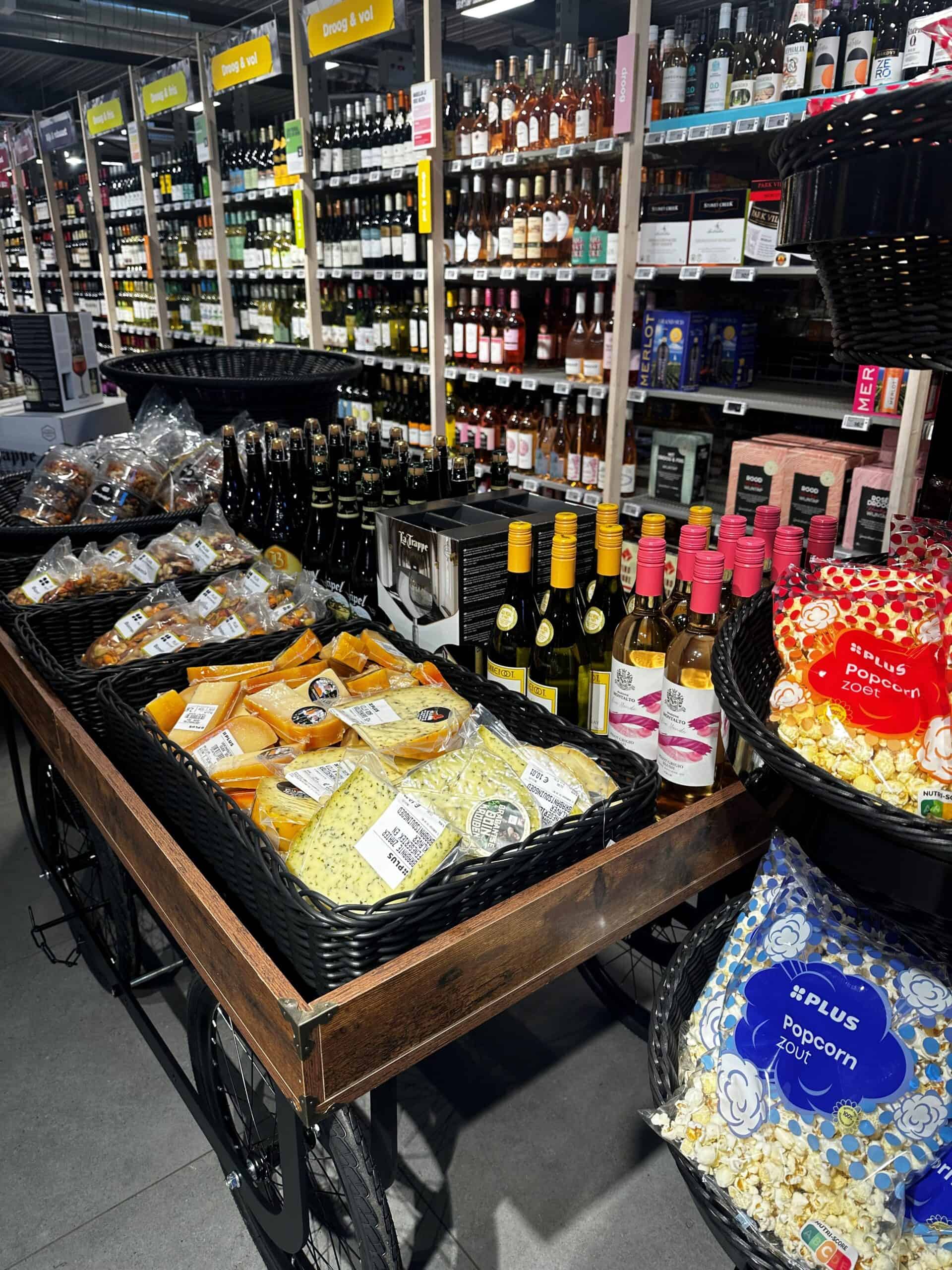 Verschillende kazen, wijn en snacks in een winkel, met wijnflessen en popcorn op een houten winkelwagen, ideaal voor wijn- of kaasliefhebbers.