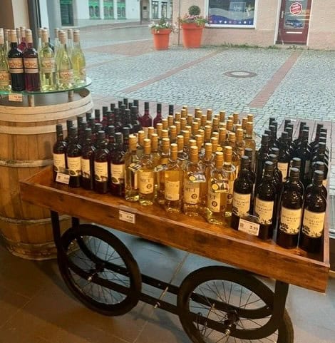 Rij met diverse wijnflessen op een houten kar, perfect voor wijnverkoop of proeverijen, binnen in een winkel met straatzicht.