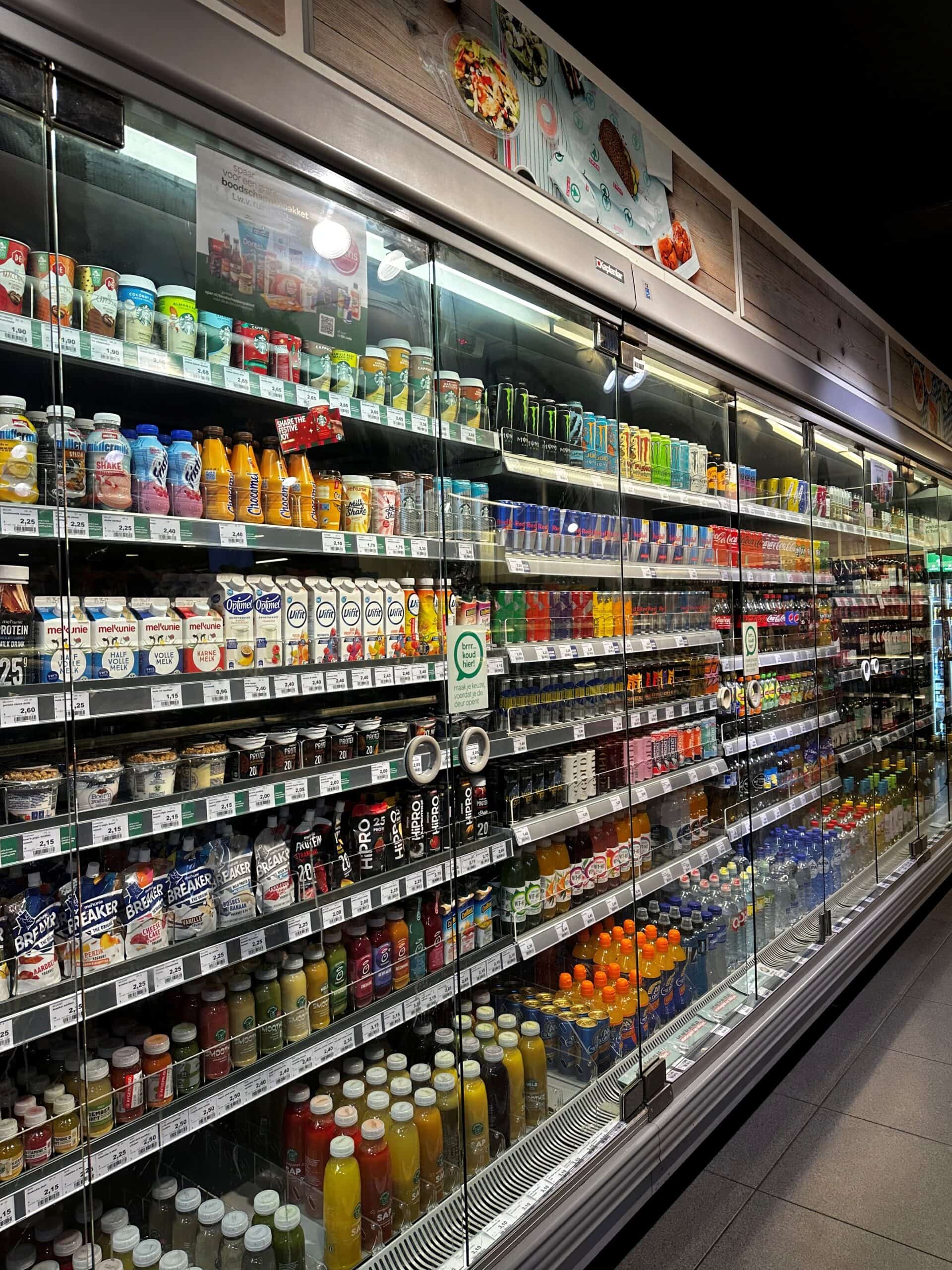 Zoekmachinevriendelijke ALT-tekst: Frisdrank en zuivelproducten in gekoelde vitrines bij supermarkt, brede selectie, inclusief energiedranken, vruchtensappen en melk.