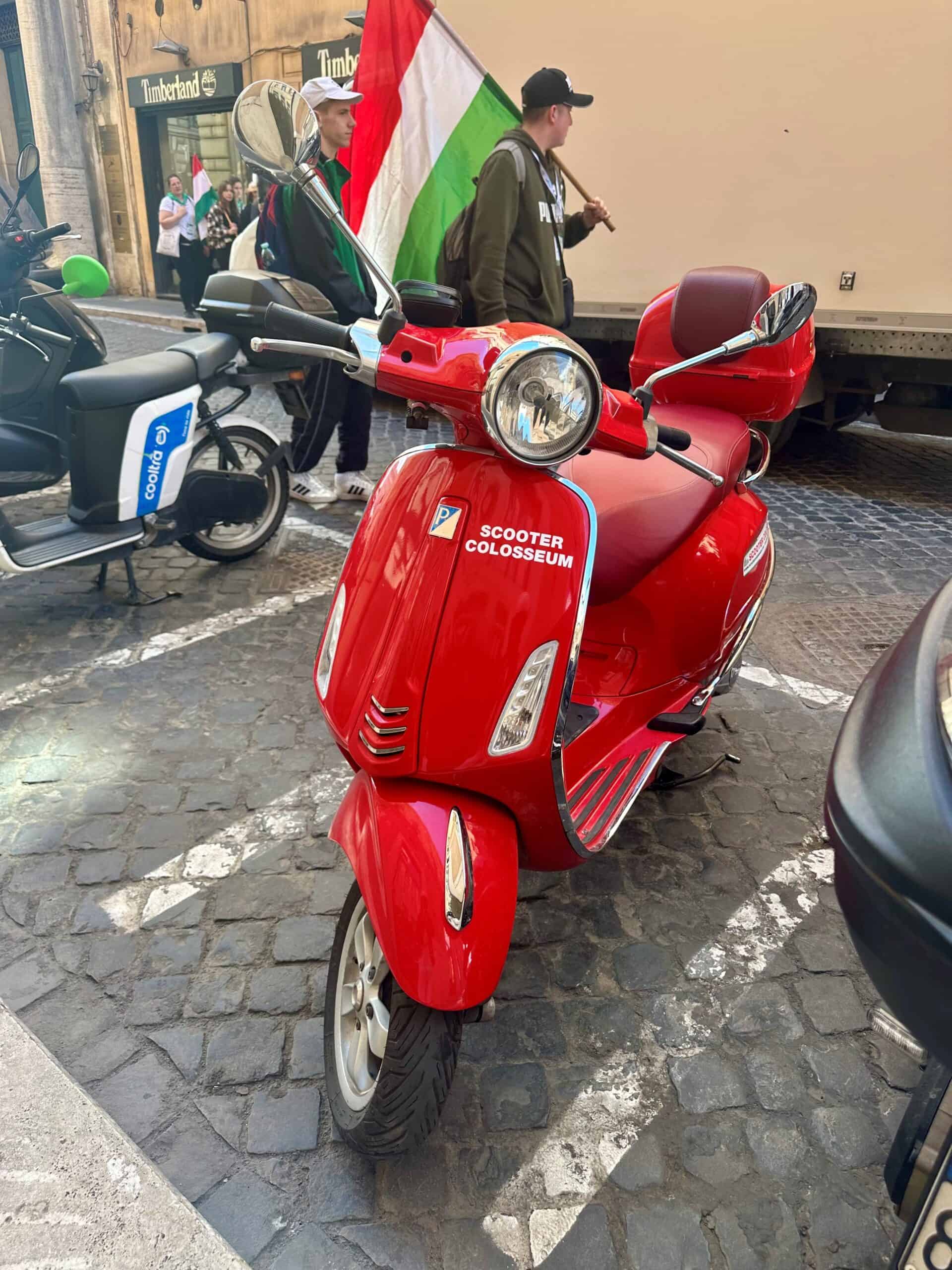 Scooter op straat met "Scooter Colosseum" sticker, omringd door andere elektrische voertuigen, straatbeelden en mensen, benadrukt modern mobiliteitsoplossingen en elektrische scooters in stedelijke omgevingen.
