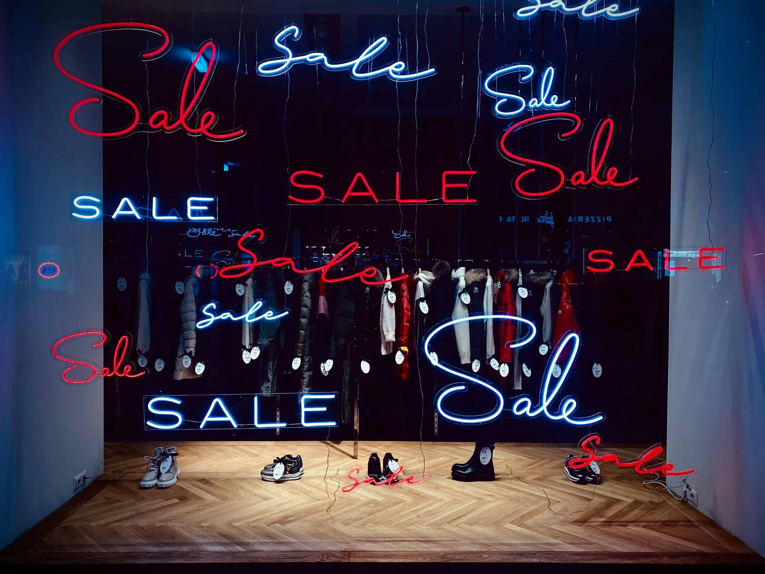 Verlichte winkelraam met neonletters die 'Sale' promoten, met kleding en schoenen in de etalage, benadrukt de commerciële en verkoopgerichte sfeer voor e-commerce en detailhandel.
